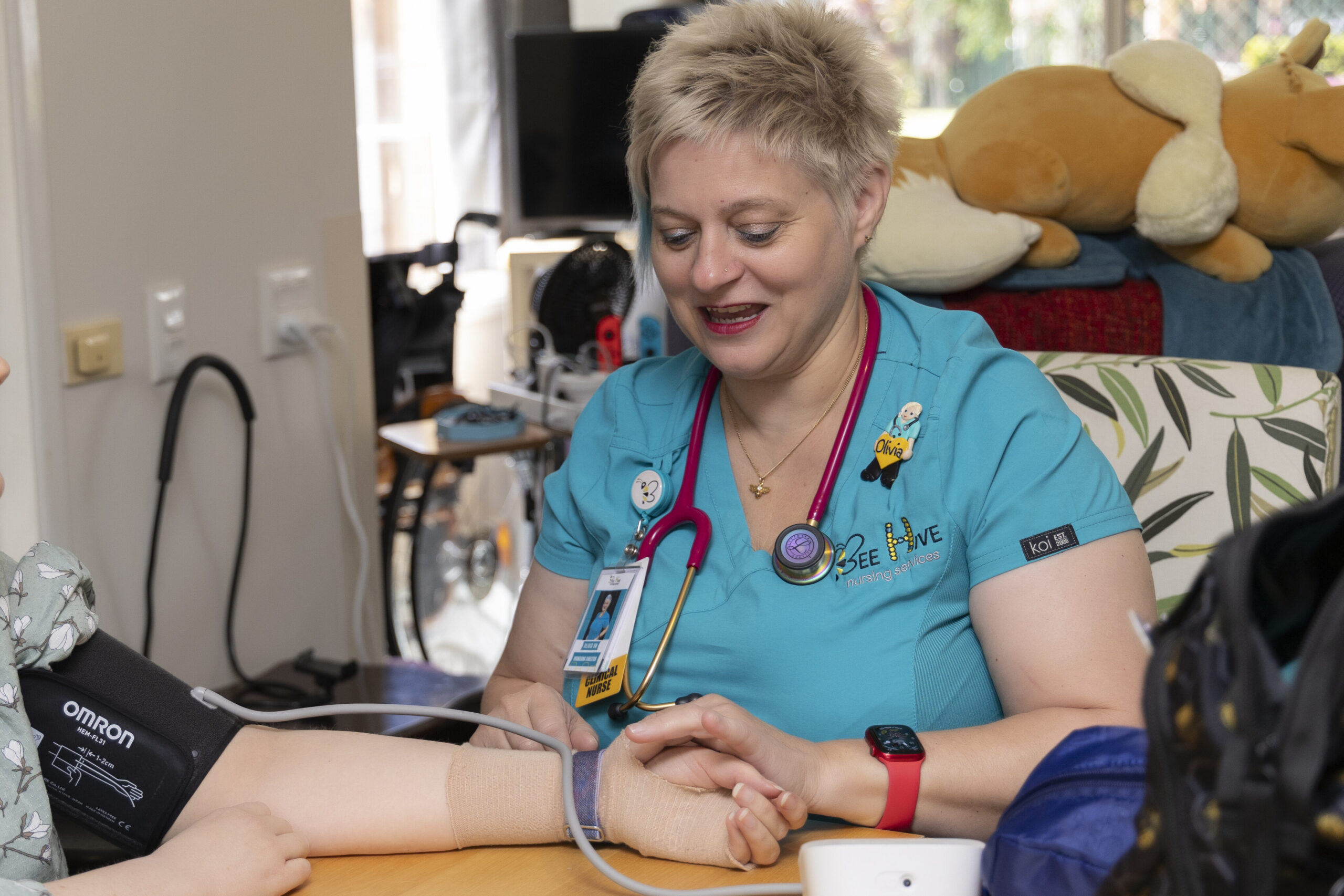 Nurse with spiky short hair wearing teal scrubs sitting at a table taking a patients  blood pressure.