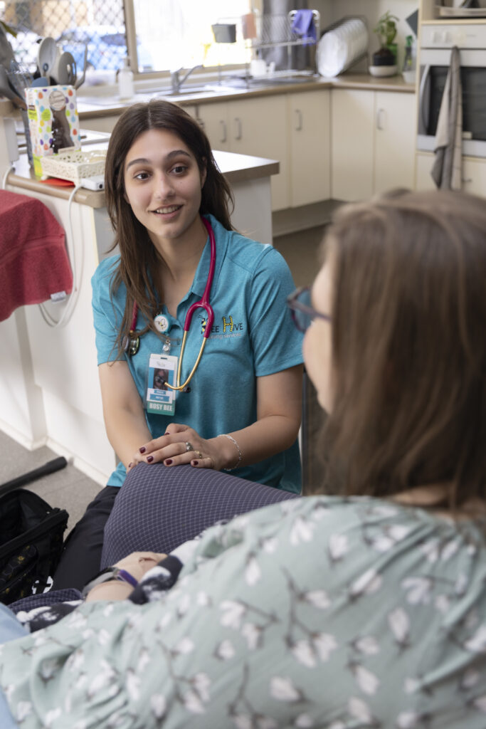 Young female nurse with dark brown ling hair in teal nurses scrubs, talking to a client who is seated in a dark blue chair with her back to the camera.